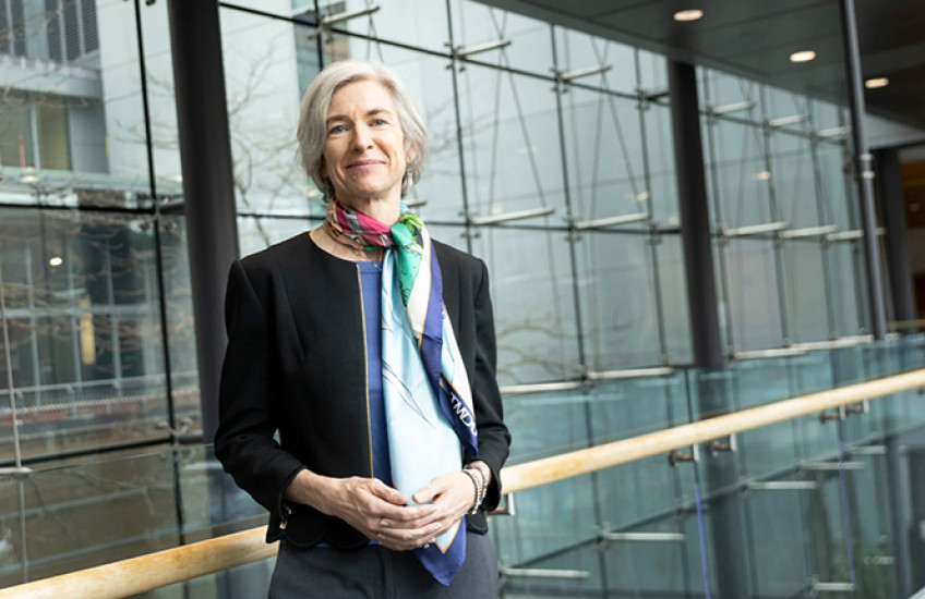 portrait photo of Jennifer Doudna, a slightly smiling white woman with chin-length gray hair, leaning against a railing in front of a wall of windows. she wears a blazer and colorful silk scarf