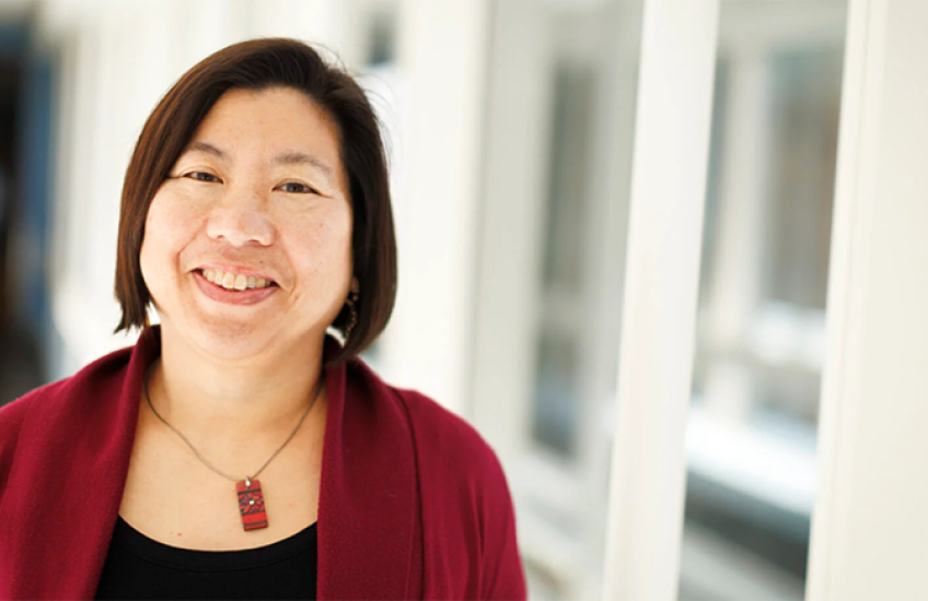 photo of a smiling asian-american woman with red blazer and necklace standing in a high-tech-looking corridor with many windows
