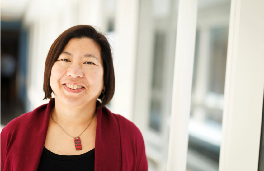 Head shot of a smiling Asian-American woman with red blazer and necklace, standing in a high-tech-looking corridor with many windows
