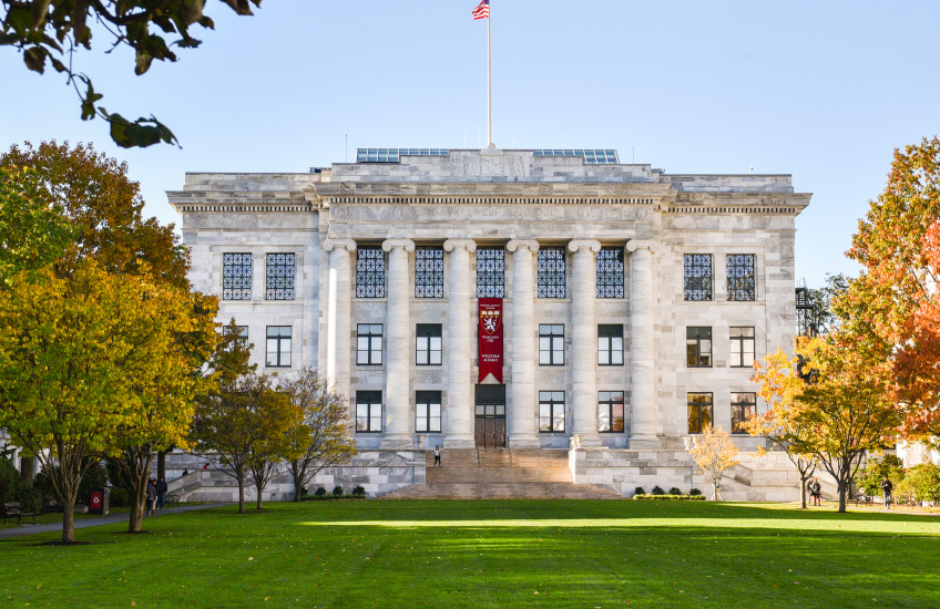 Colorful photograph of Gordon Hall on the HMS campus, a marble building with columns. In front is a manicured lawn and autumnal trees