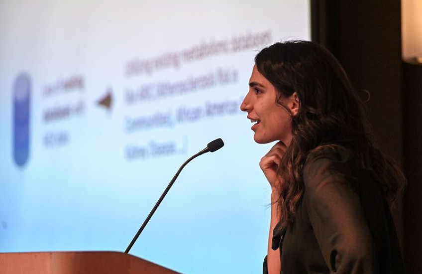 A young woman seen from the side presents at a lectern in front of a projected slide show