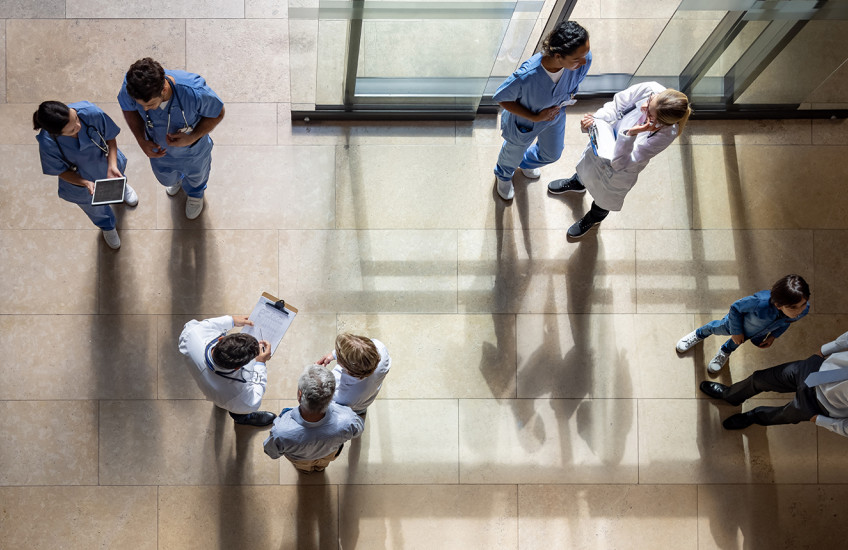 An overhead view of nine people in the entryway of a hospital, standing in groups of two or three, many dressed in medical gear or lab coats, carrying clipboards and digital devices.