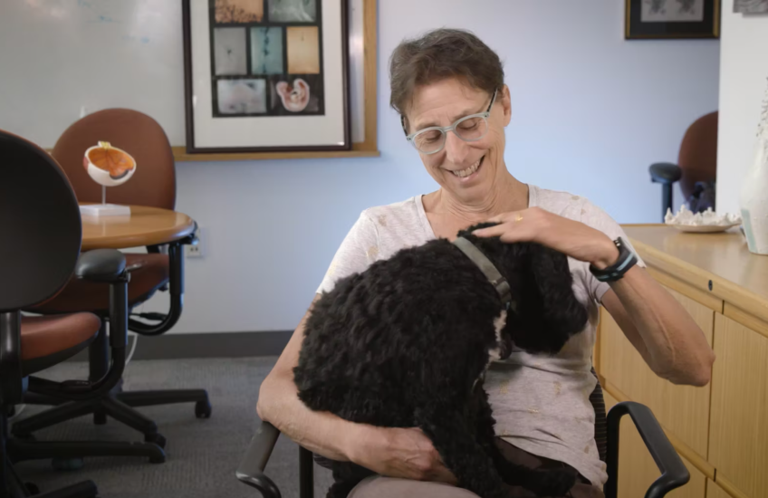 Screen shot of a woman with short hair smiling down at a cockapoo dog sitting in her lap. She is in an office with a table and chairs and framed art in the background.