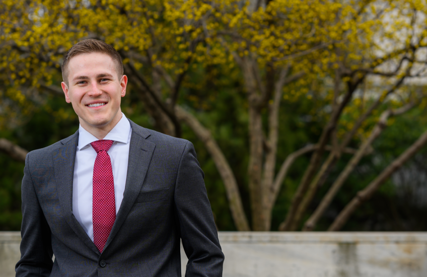 Winkie in blue suit and crimson tie standing in front of a bench on Quad with budding tree behind him
