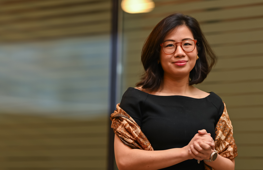 A woman in a black dress and colorful shawl stands in front of a window.