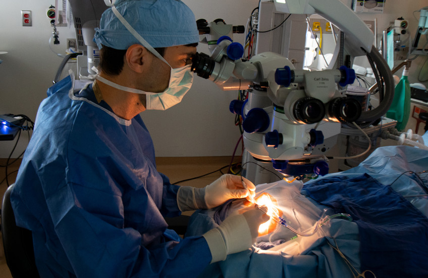 Wearing tanslucent white gloves and a blue cap, mask, and medical garb, a surgreon manipulates several objects connected to wires and tubes above the patient's eye. The patient's face is invisible in the intense glow of the operating lights.