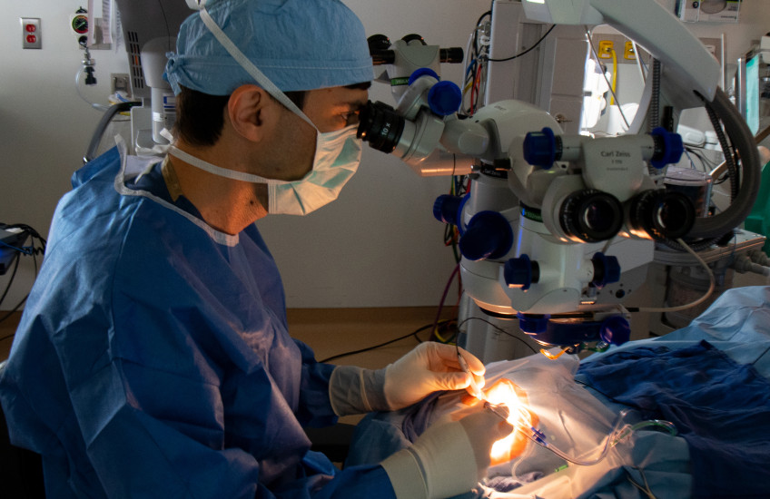 Wearing tanslucent white gloves and a blue cap, mask, and medical garb, a surgreon manipulates several objects connected to wires and tubes above the patient's eye. The patient's face is invisible in the intense glow of the operating lights.