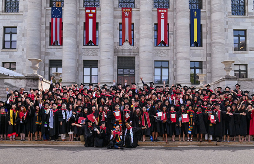 Wide-angle photograph of MD and DMD graduates in regalia cheering on the front steps of Gordon Hall, five tall flags hung between marble columns behind them