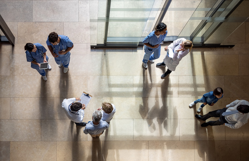 An overhead view of nine people in the entryway of a hospital, standing in groups of two or three, many dressed in medical gear or lab coats, carrying clipboards and digital devices.