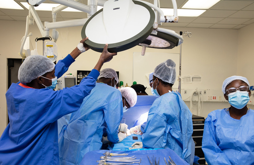 A surgical team of doctors and nurses wearing blue scrubs, gauzy head coverings, and medical masks adjust lights, provide anesthesia, and perform other actions necessary for an operation, all gathered around the patient on the operating table. Health equity requires access to surgical care.