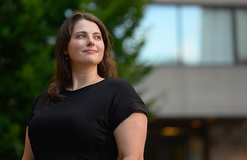 A young woman in a black short sleeve dress stands outside smiling with a building and tree in the background