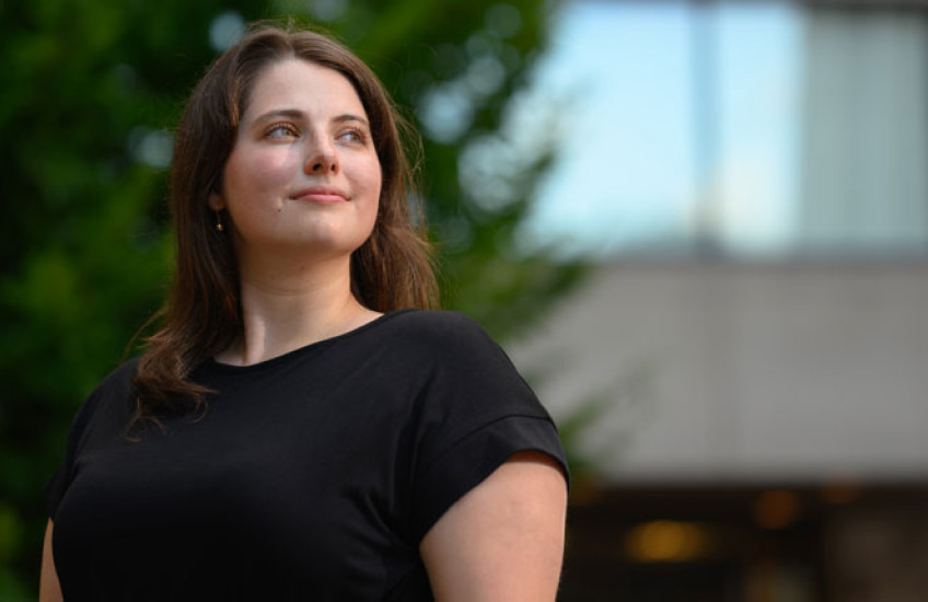 A young woman in a black short sleeve dress stands outside smiling with a building and tree in the background