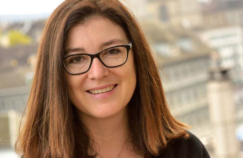 Portrait photo of a smiling woman with glasses and shoulder-length hair, sitting on a wall with a city skyline out of focus in the background.