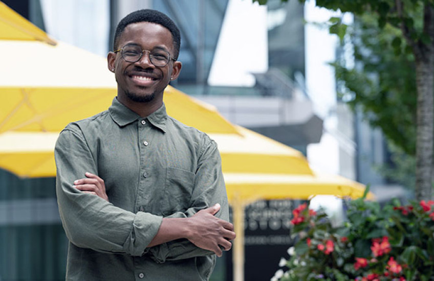A smiling young Black man with glasses stands with arms crossed in front of picnic tables on the HMS Quad