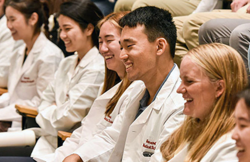 Seven medical and dental students wearing white coats seated in amphitheater smiling and laughing as they listen to patients.