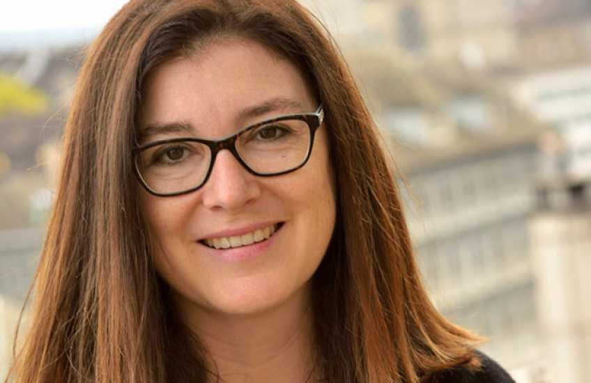 Portrait photo of a smiling woman with glasses and shoulder-length hair, sitting on a wall with a city skyline out of focus in the background. 
