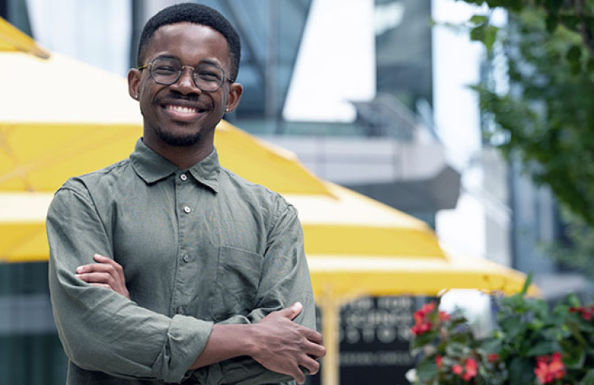 A smiling young Black man with glasses stands with arms crossed in front of picnic tables on the HMS Quad