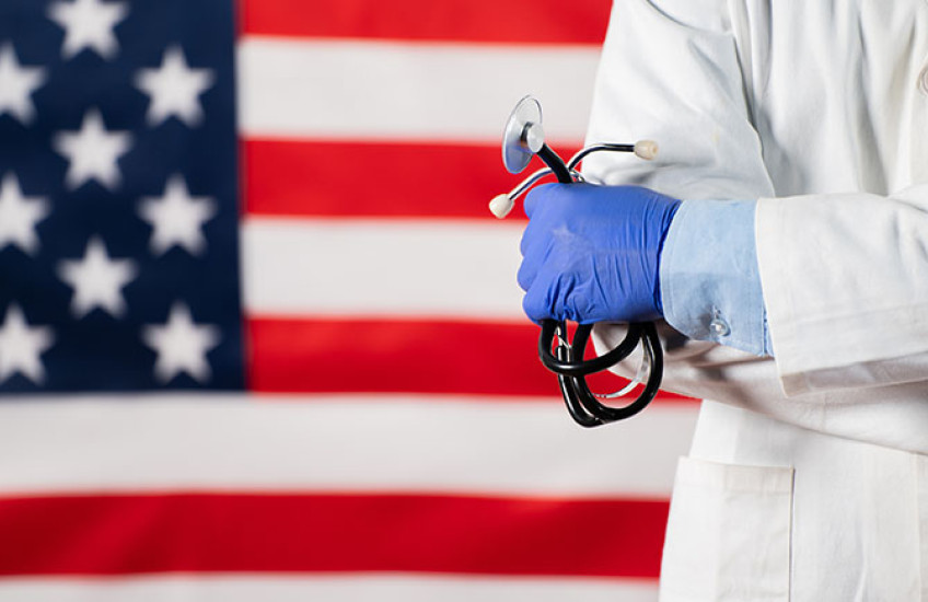 A health care practitioner wearing a white coat and holding a stethoscope stands in front of an American flag