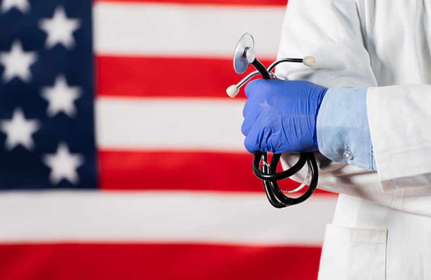 A health care practitioner wearing a white coat and holding a stethoscope stands in front of an American flag