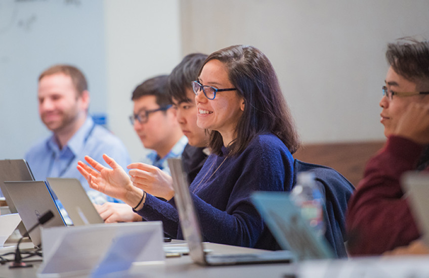 Six trainees at a conference table with laptops in front of them. The one at center speaks with a smile and gesturing hands.