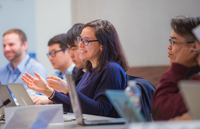 Six trainees at a conference table with laptops in front of them. The one at center speaks with a smile and gesturing hands.