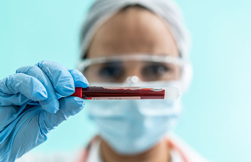 A person wearing protective gloves, glasses, and mask holds a small vial containing a blood sample.