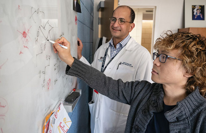 Two scientists — one in a lab coat and one in a cardigan — collaborate at a whiteboard with chemical diagrams