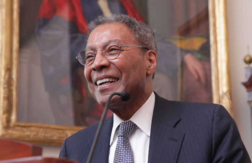 A man in a suit smiles at a lectern in front of a painting of someone in academic robes