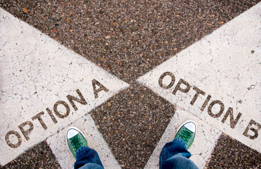 An aerial view of two feet in green sneakers, one standing on a large white arrow labeled “option A” and the other standing on a large white arrow labeled “option B”
