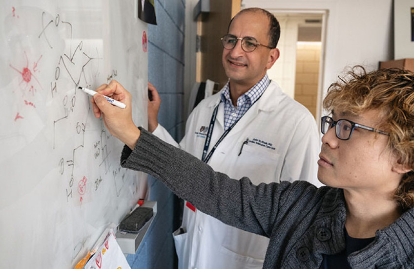 Two scientists — one in a lab coat and one in a cardigan — collaborate at a whiteboard with chemical diagrams