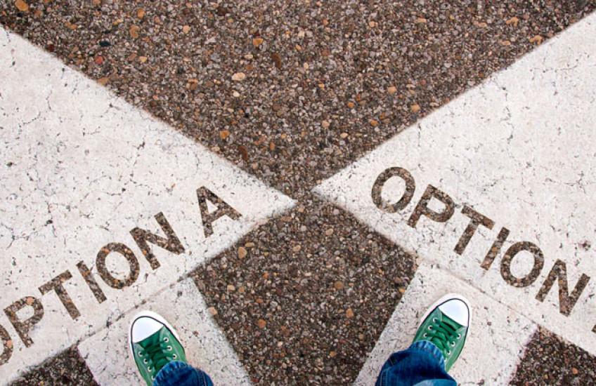 An aerial view of two feet in green sneakers, one standing on a large white arrow labeled “option A” and the other standing on a large white arrow labeled “option B”