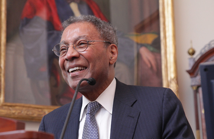 A man in a suit smiles at a lectern in front of a painting of someone in academic robes