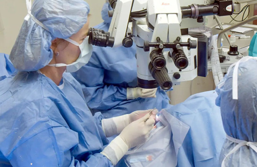In an operating room, three people in blue surgical gowns, masks, and head covers look through microscopes while performing surgery on a patient.