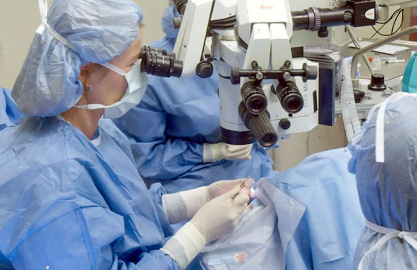 In an operating room, three people in blue surgical gowns, masks, and head covers look through microscopes while performing surgery on a patient.