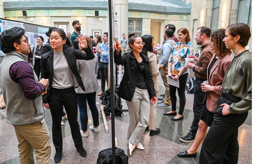 Two students on either side of a row of posters present their work to numerous onlookers