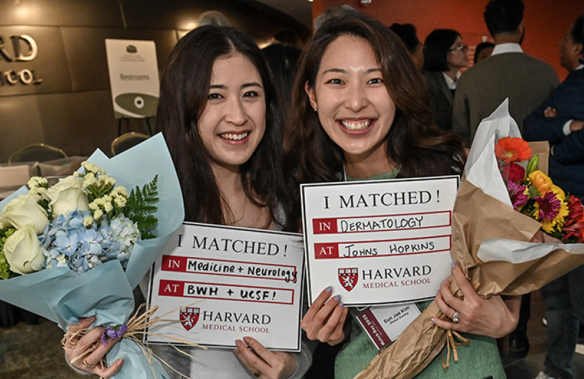 Two smiling women hold bouquets and Match Day signs from Harvard Medical School, celebrating their residency placements in Medicine + Neurology and Dermatology.