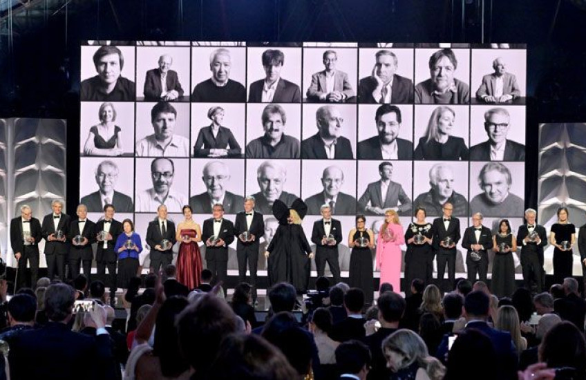 A row of Breakthrough laureates standing on a stage with black and white headshots of previous winners projected in the background