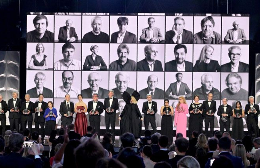 A row of Breakthrough laureates standing on a stage with black and white headshots of previous winners projected in the background