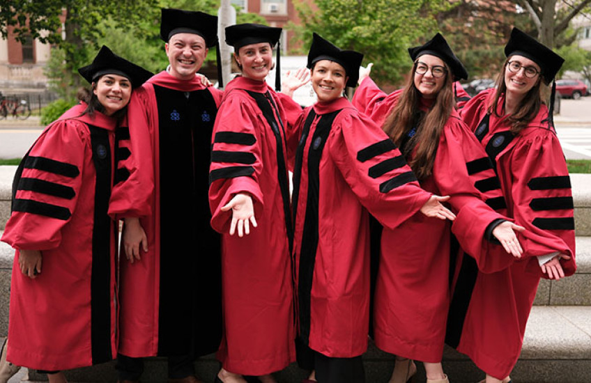 Five women and one man in academic regalia smile and stretch out their hands, tah dah.