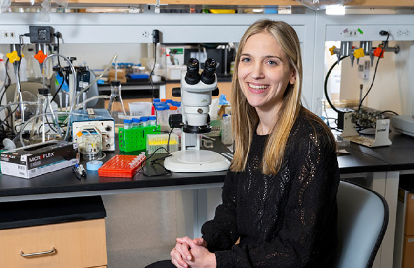 A researcher sits on a chair next to a microscope in a lab space