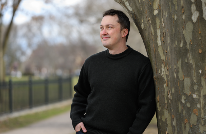 A man leans against a tree, smiling and looking off into the distance