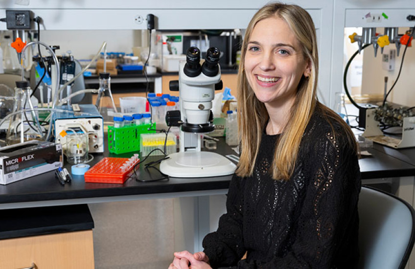 A researcher sits on a chair next to a microscope in a lab space