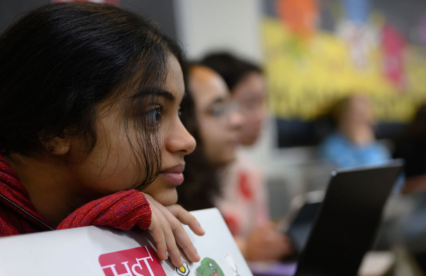 Focus on one student leaning on laptop with an HST sticker on the cover with several students in blurred background.