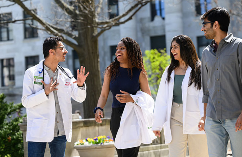 Four medical students, two in white coats, smiling and walking together on a sunny day with a marble columned building in background.