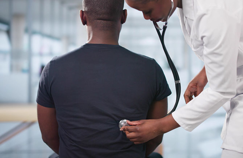 A doctor in a white lab coat uses a stethoscope to listen to the back of a seated patient wearing a dark t-shirt, in a bright, modern medical office.