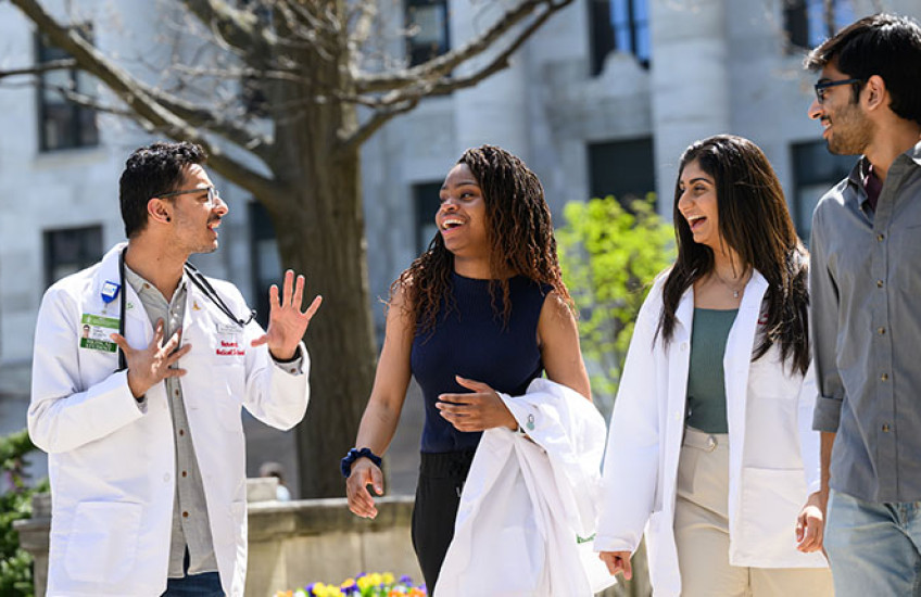 Four medical students, two in white coats, smiling and walking together on a sunny day with a marble columned building in background.