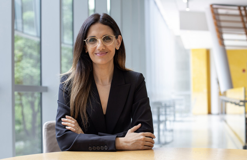 A woman with long, straight hair and glasses sits at a round wooden table in a modern, sunlit building with large windows. She is wearing a black blazer and smiling confidently with arms crossed.
