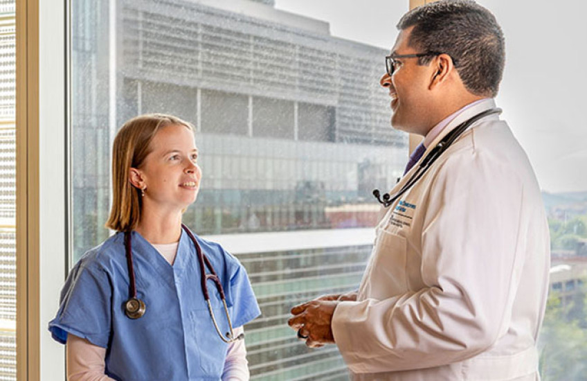A woman in blue scrubs and a man in a white coat, both wearing stethoscopes, stand talking to each other in front of a window.