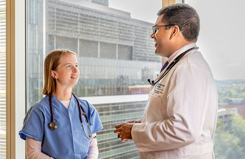 A woman in blue scrubs and a man in a white coat, both wearing stethoscopes, stand talking to each other in front of a window.