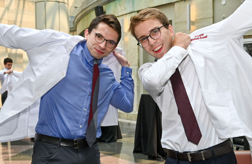 Two men put on white coats in a sun-filled atrium.